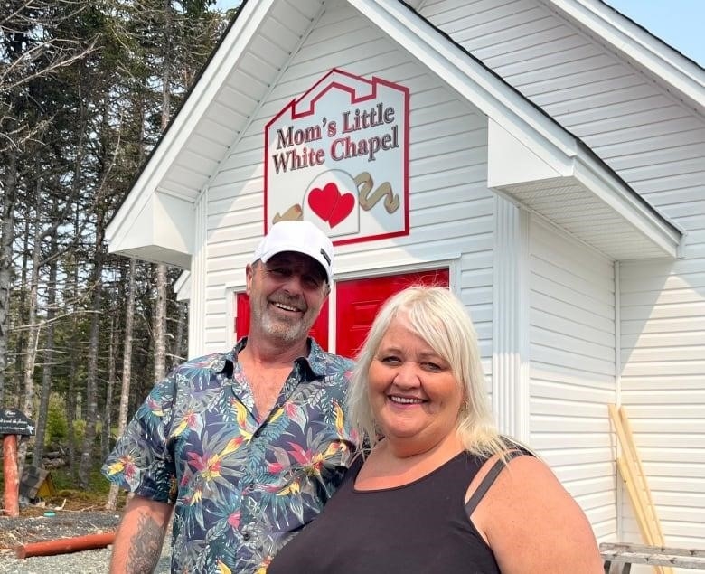 A man, left, and a woman smile. They stand in front of a chapel. A sign above the door reads "Mom's Little White Chapel".