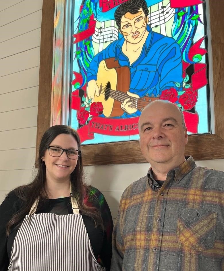 A woman and a man stand next to each other and smile. Behind them is a window depicting Elvis with a guitar in his hands.