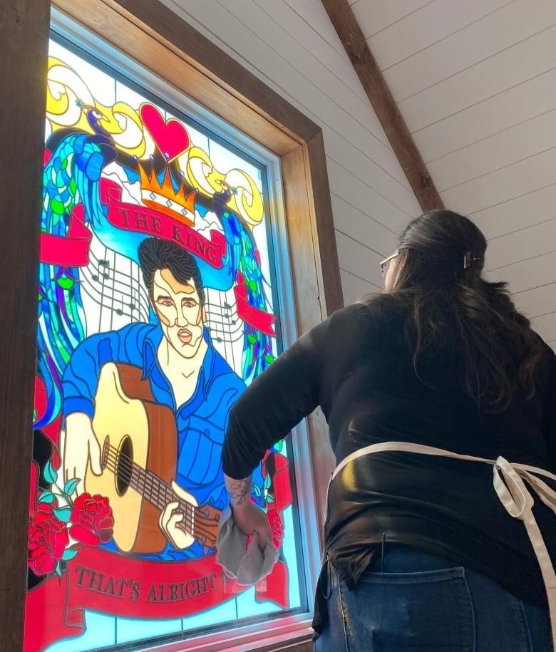 A woman stands in front of a window that shows Elvis and wipes it with a cloth.